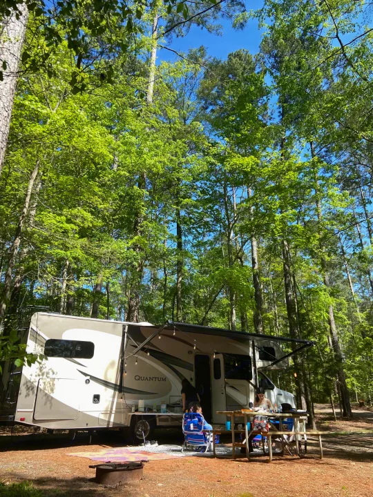 An RV rental (Quantum) at a sunny campsite with people relaxing at a picnic table under string lights amidst tall green trees.. Thor Quantum 2017