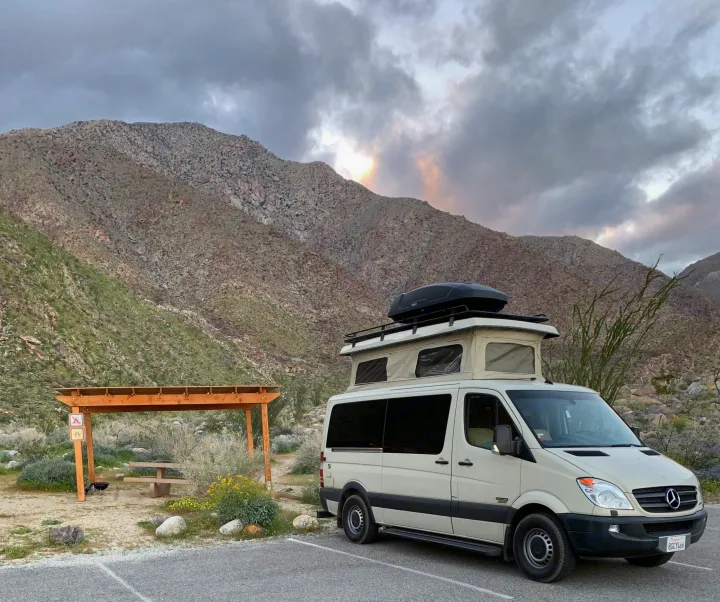 Beige RV rental camper van parked at a desert mountain campsite with a pop-top roof and cargo box, picnic area nearby.. Sportsmobile Regular Body 2013