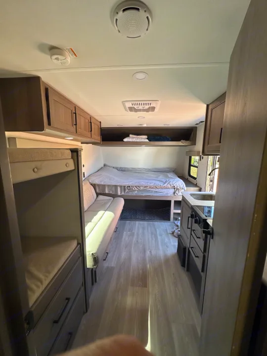 Bright RV rental interior showcasing bunk beds on the left, a main bed, and a compact kitchen counter on the right.. Transcend One 2025 2025