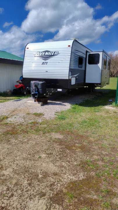Avenger travel trailer RV rental with a slide-out deployed, parked on a gravel lot beside a building. An ATV is near.. Avenger ATI 2019