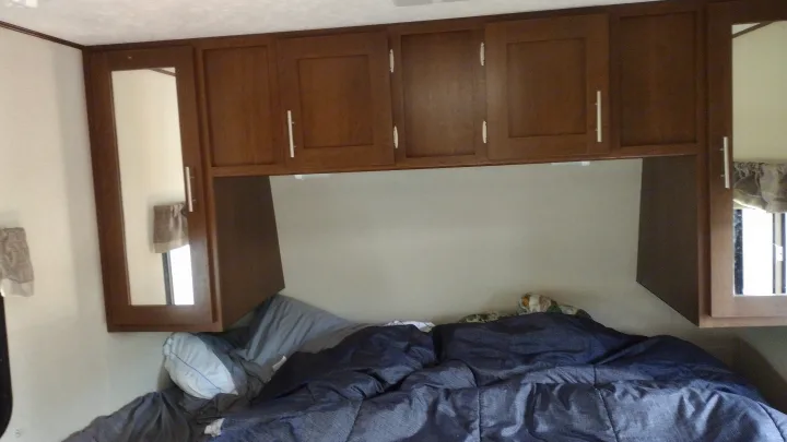 Inside an RV rental bedroom with wooden overhead cabinets, mirrored side cabinets, and a comfy bed with blue bedding.. Avenger ATI 2019