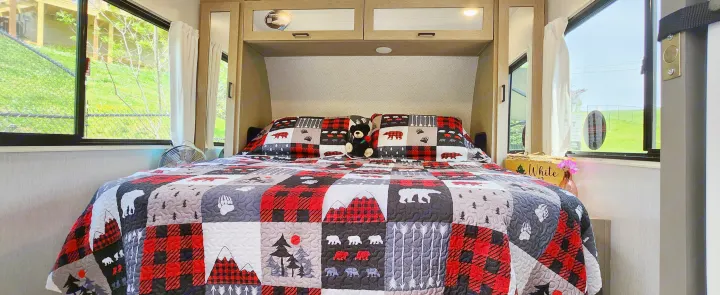 Cozy RV rental bedroom with a bear-themed quilt, overhead cabinets, and scenic windows. Perfect for your next adventure!. Forest River Wildwood Platinum 2024