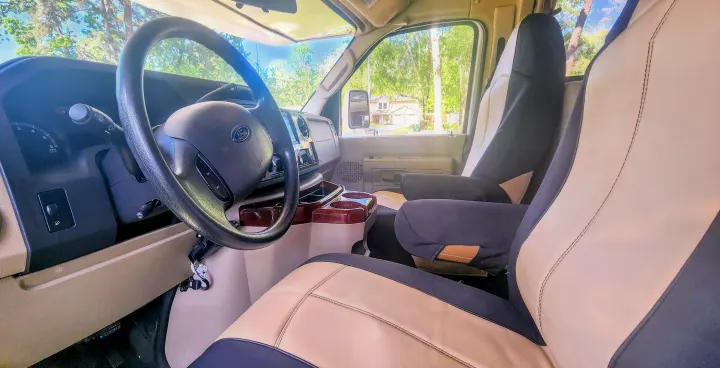 Detailed view inside an rv rental: driver's area with Ford steering wheel, dashboard, and comfortable black/beige front seats.. Forest River Coachmen Leprechaun 2015