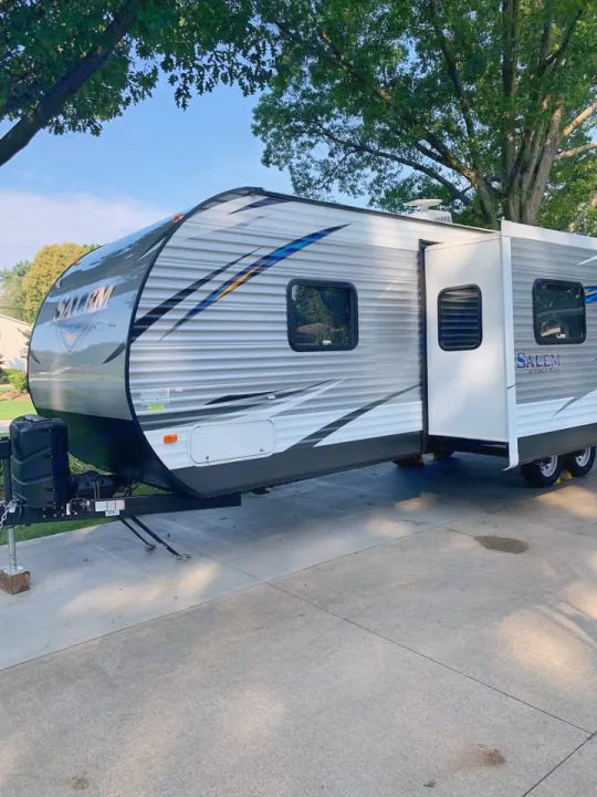 This silver and white Salem RV travel trailer, perfect for an RV rental, is parked on a driveway with its slide-out extended.. Forest River Salem 2018