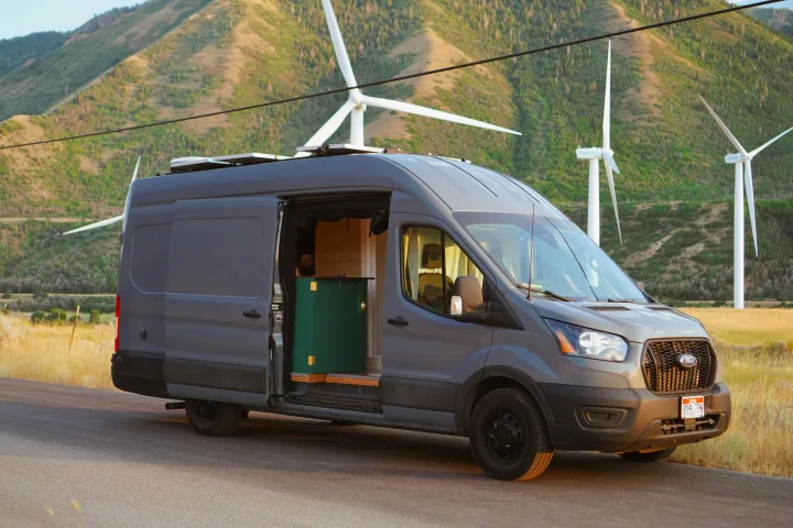 Gray Ford Transit camper van with solar panels, ideal for an rv rental, parked near wind turbines on a scenic hillside.. Ford Transit Custom 2022