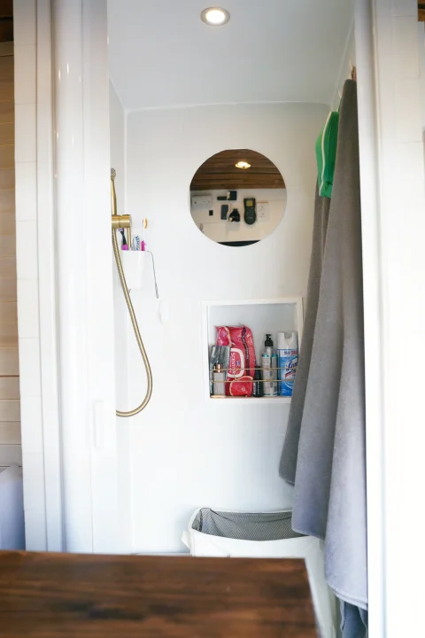 Inside an RV rental bathroom: shower, shelf with cleaning supplies, mirror, and laundry hamper for compact living.. Ford Transit Custom 2022