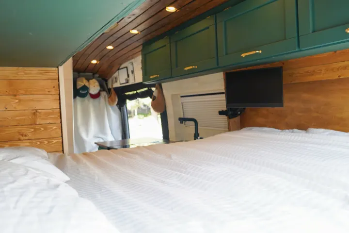 Spacious RV rental interior. Features a bright bed, green overhead cabinets, wood paneling, and a TV. Natural light from a window.. Ford Transit Custom 2022