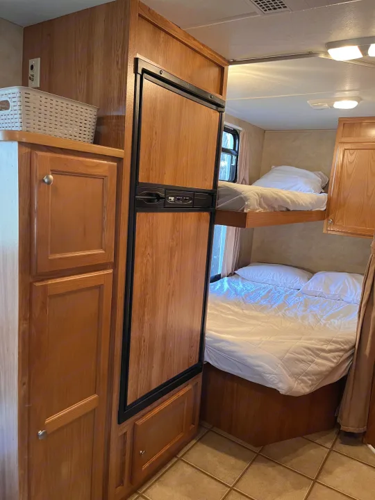 RV rental interior: wood-paneled fridge, bunk beds with white bedding, cabinets, and tiled floor.. R-Vision Trail Sport 2014