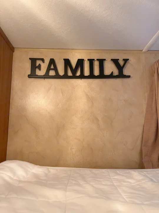 Black 'FAMILY' sign with hooks on a textured tan wall above white bedding in an RV rental.. R-Vision Trail Sport 2014