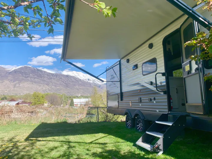 Enjoy stunning views from this RV rental! A camper sits on green grass with an extended awning, facing snowy mountains.. Jayco Jay Flight 2024