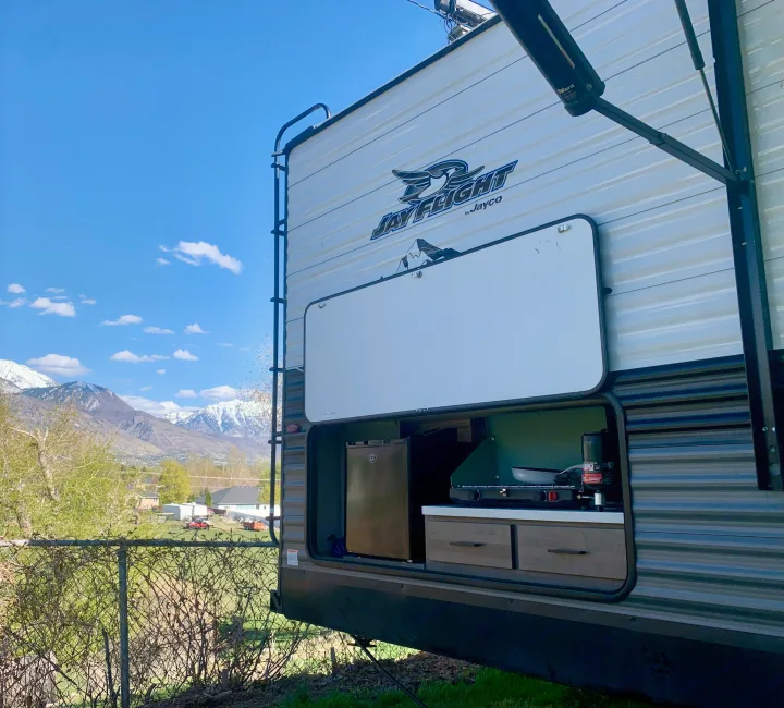 This Jayco Jay Flight rv rental features an open outdoor kitchen with a fridge and stove, set against snowy mountains.. Jayco Jay Flight 2024