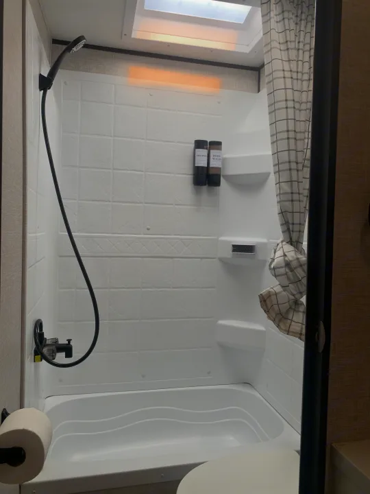 Modern RV rental bathroom featuring a white shower/tub combo, black shower head, and wall-mounted dispensers. Plaid curtain.. Jayco Jay Flight 2024