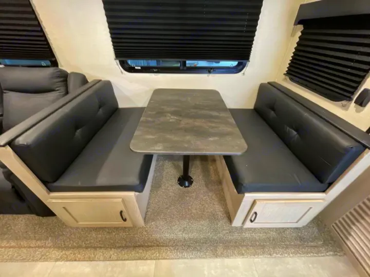 This RV rental dinette features black cushioned booth seating with storage, a gray table, and window blinds. Ideal for travel.. Coachmen Apex 2022