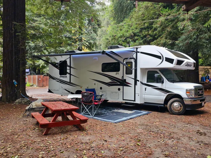 A white Jayco RV rental is parked at a shaded forest campsite, featuring a picnic table, chairs, and an awning.. Jayco Redhawk 2024