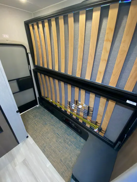 RV rental interior features a folded-up bunk bed with visible slats, a textured floor, and an exit door, optimizing space.. Ford Transit 2023