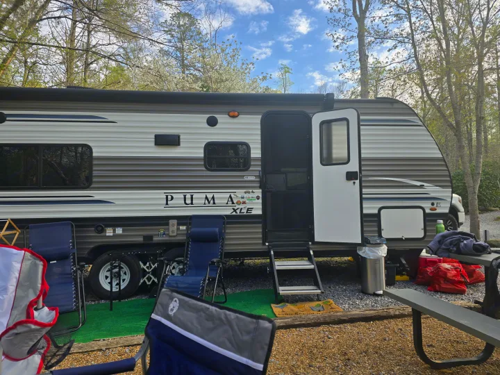 An rv rental (Puma XLE) is parked at a serene campsite, complete with chairs, picnic table, and outdoor mat.. Palomino Puma 2022