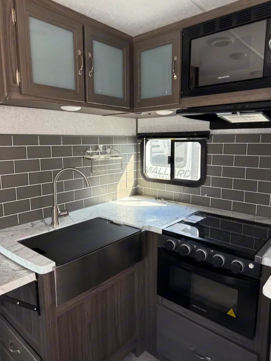 Modern RV rental kitchen with dark wood cabinets, gray subway tiles, marble-look counters, sink, oven, and microwave.. Dutchmen Kodiak Ultra Lite 2021