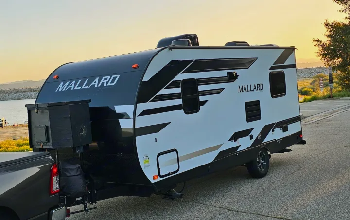 White and black Mallard travel trailer, perfect for rv rental. Hitched to a truck, parked by a scenic lake at sunset.. Heartland RVs Mallard 2021