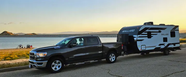 RV rental adventure: A Ram truck tows a Mallard travel trailer by a scenic lake with sunset-lit mountains and distant campers.. Heartland RVs Mallard 2021