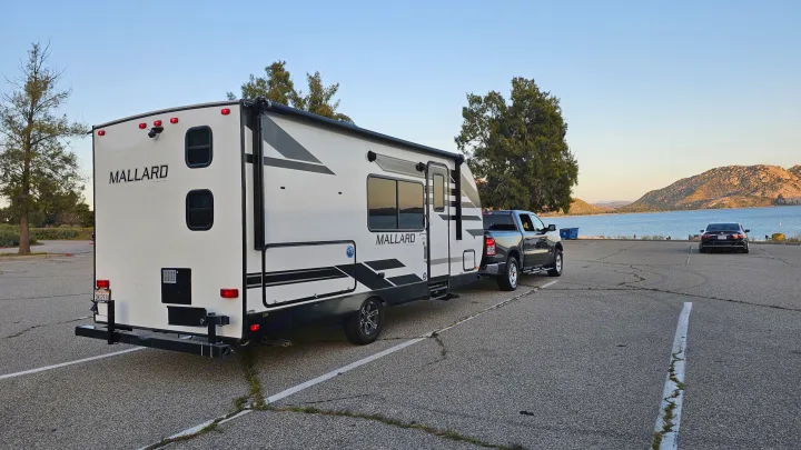 A white Mallard travel trailer RV rental, hitched to a pickup truck, parked near a scenic lake with mountains.. Heartland RVs Mallard 2021
