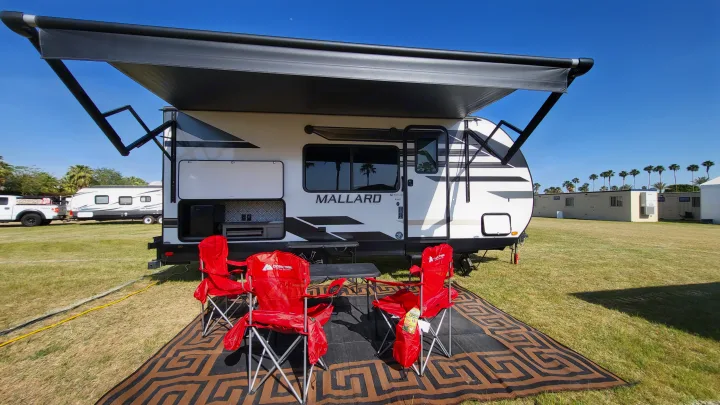 White Mallard RV rental with extended awning, outdoor kitchen, red chairs on a patterned rug for sunny camping.. Heartland RVs Mallard 2021