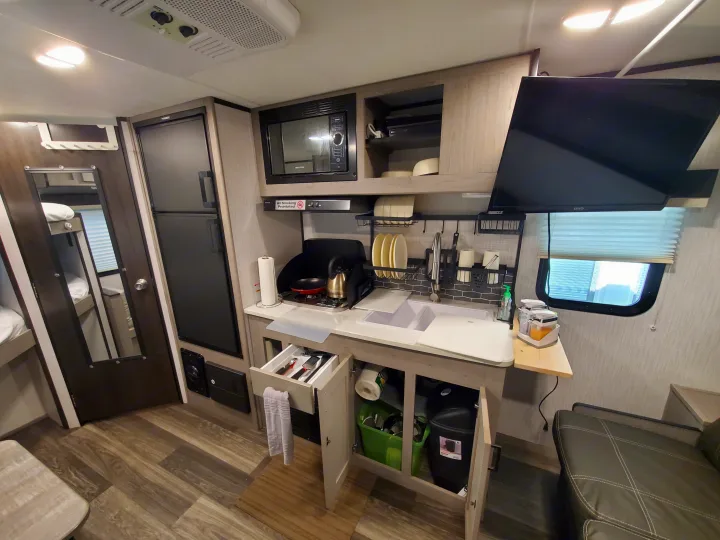 Detailed view of an RV rental's kitchen, showcasing a sink, microwave, fridge, stove, and a glimpse of the living area with a TV.. Heartland RVs Mallard 2021