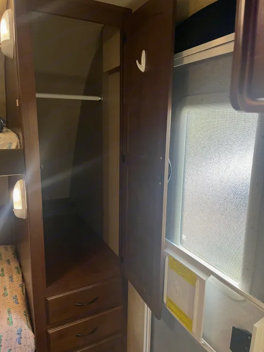 RV rental interior featuring an open wardrobe with a hanging rod and drawers, alongside a bunk bed and a screened window.. Jayco Jay Feather Ultra Lite 2014