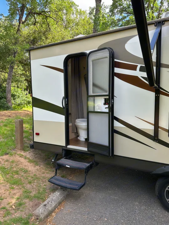 RV rental showing its open door with a compact bathroom & toilet inside, parked at a leafy campsite.. Jayco White Hawk Ultra Lite 2015