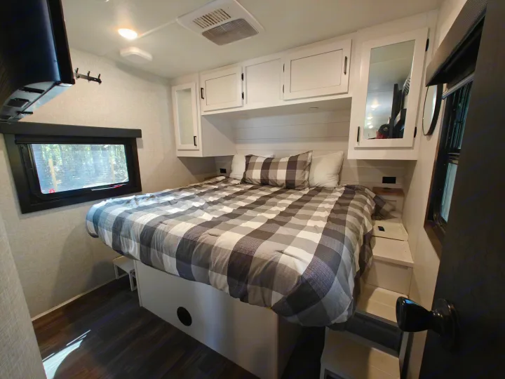 This RV rental bedroom features a cozy plaid bed, modern white cabinets, and a window view. Perfect for your next adventure!. Highland Ridge Open Range Light 2024
