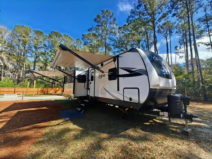 An RV rental with awnings extended, parked in a wooded campsite with pine trees and a clear blue sky.. Highland Ridge Open Range Light 2024