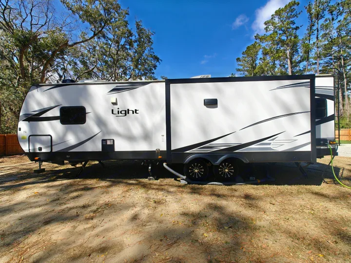 White "Light" travel trailer RV for rv rental, parked and hooked up at a sunny treed campsite.. Highland Ridge Open Range Light 2024