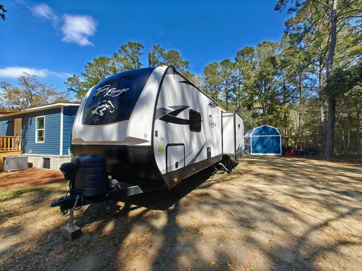 Open Range Light RV rental parked on a sunny, dirt lot near a blue house & shed, surrounded by trees.. Highland Ridge Open Range Light 2024