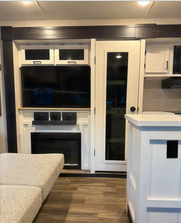 Modern rv rental interior: entertainment center with TV/fireplace, light sofa, glass door, and white kitchen island with outlet.. Highland Ridge Open Range Light 2024