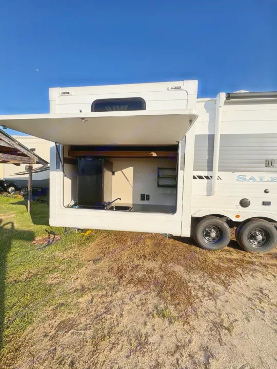 This white RV rental features an open outdoor kitchen with a fridge, sink, and counter space, parked on grass/dirt under a blue sky.. Forest River Salem 31kqbts 2025