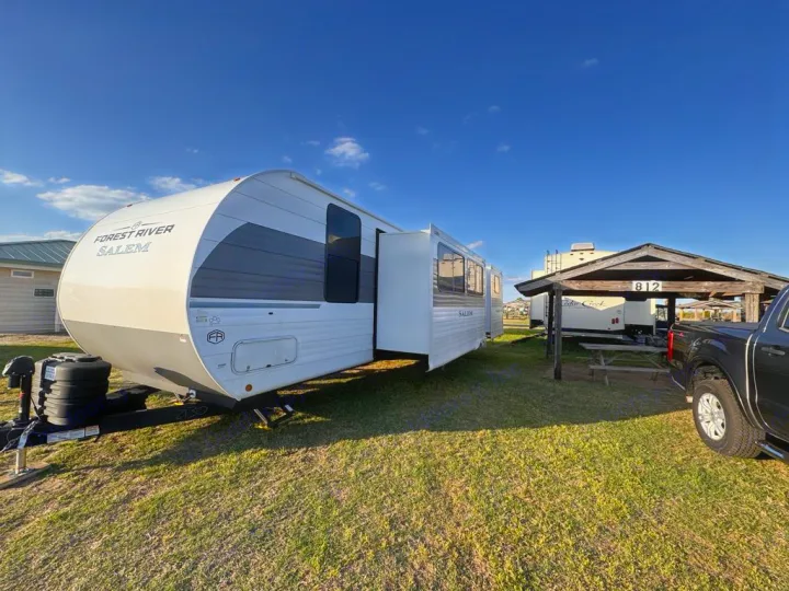 Forest River Salem travel trailer RV rental, white with gray accents, parked on grass. Another RV and truck visible under a shelter.. Forest River Salem 31kqbts 2025