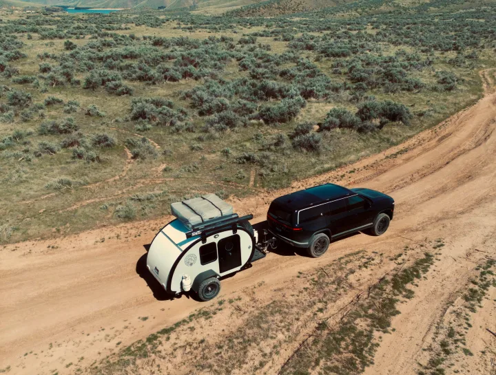 Black Rivian SUV towing a white teardrop camper on a dirt road. Explore nature with this ideal RV rental setup.. Bean Trailer Bean Trailer Trailer 2019