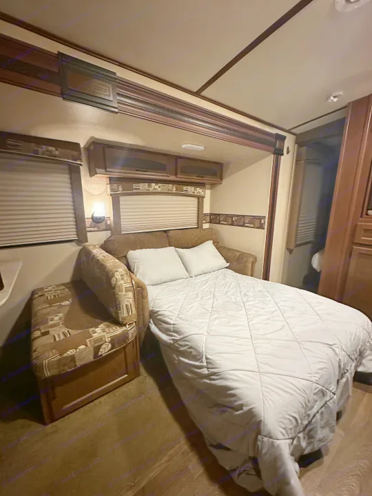 Cozy RV rental interior. A sofa bed, extended & covered by a light comforter, with dark wood cabinetry & blinds.. Jayco White Hawk Ultra Lite 2015