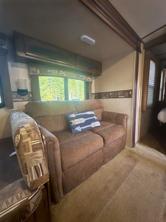 View inside an RV rental showing a comfortable brown sofa with a pillow, a window with trees, and carpeted floor.. Jayco White Hawk Ultra Lite 2015