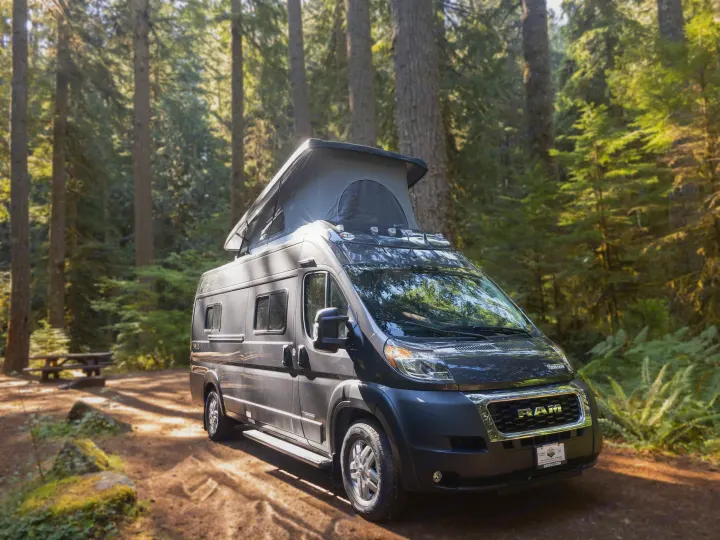 Dark grey pop-top camper van for rv rental parked in a sunny forest. Ideal for outdoor adventures & nature camping.. Winnebago Solis 2023
