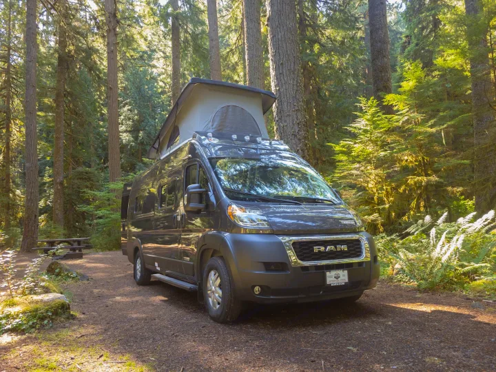 A dark grey pop-top RV rental van is parked in a beautiful, sun-dappled forest setting, ready for adventure.. Winnebago Solis 2023