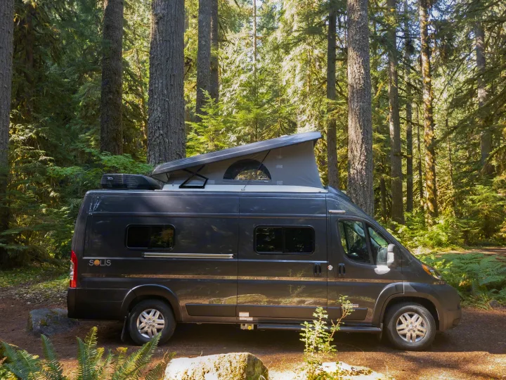Dark gray Solis camper van with pop-top extended, parked in a sun-dappled forest. Book your rv rental for nature escapes!. Winnebago Solis 2023