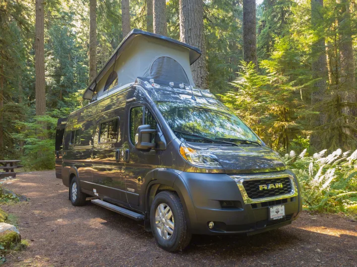A dark camper van with a pop-top roof is parked on a dirt path at a sunny forest campsite, ready for your RV rental trip.. Winnebago Solis 2023