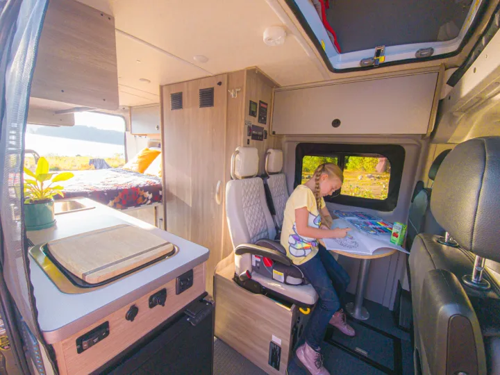 Inside a bright RV rental, a girl colors at a dinette table. A kitchen counter & bed are also visible.. Winnebago Solis 2023