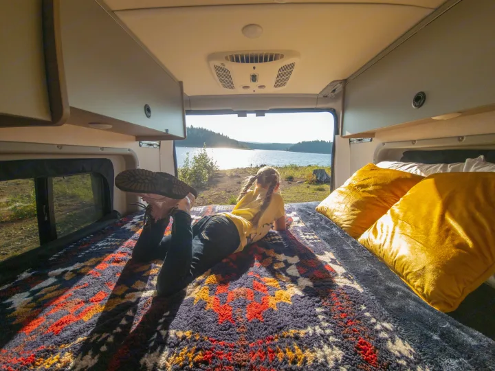 RV rental: A person lies on a colorful bed, gazing at a bright, scenic lake view from inside the camper van.. Winnebago Solis 2023