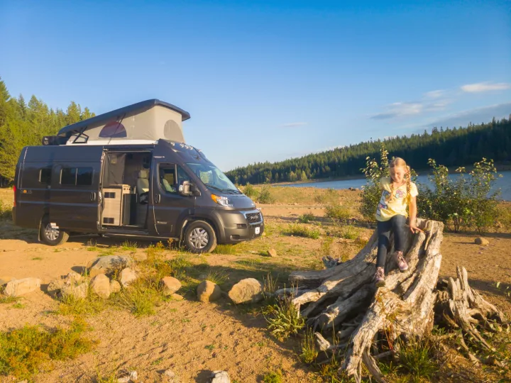 Enjoy nature with an RV rental! A young girl sits on a tree stump by a lake, next to a parked camper van with its door open.. Winnebago Solis 2023