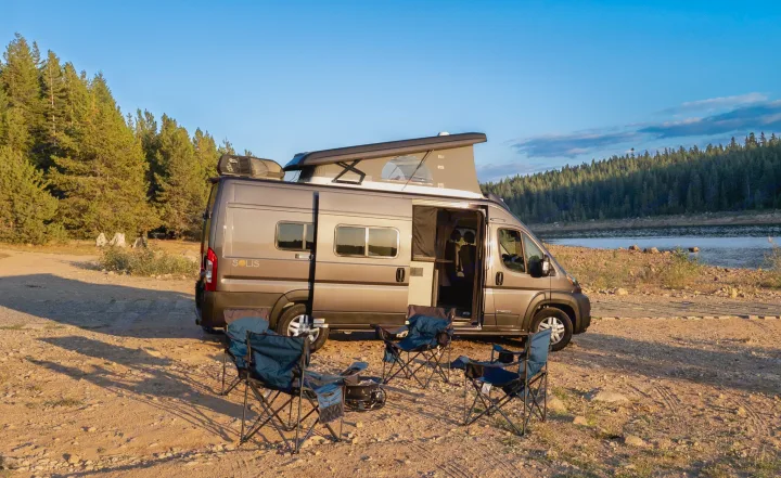 This RV rental shows a camper van ready for lakeside adventure with a pop-top, chairs, and a fire pit.. Winnebago Solis 2023