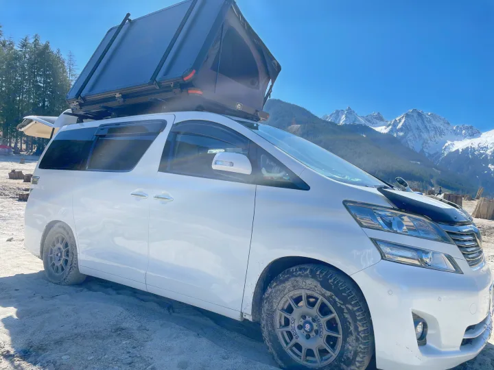 White minivan with rooftop tent, an RV rental, parked on dirt with snowy mountains and trees behind.. Toyota Dreamseeker Voyager 2008