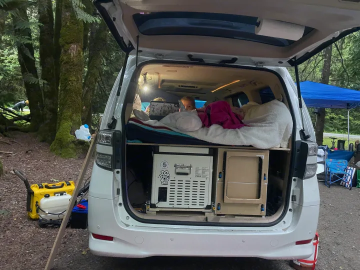 Custom minivan setup as an RV rental alternative for camping. A child is nestled in the back bed, with fridge & power below. Forest camp.. Toyota Dreamseeker Voyager 2008