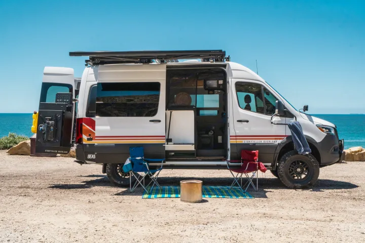 Storyteller Overland RV rental camper van parked beachside, doors open. Two chairs & fire pit set for outdoor comfort.. Storyteller Overland Storyteller Overland Class B 2022