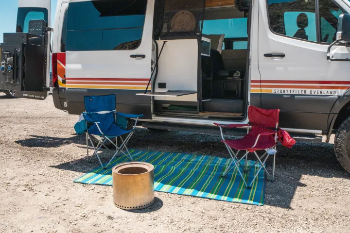 A Storyteller Overland camper van, perfect for RV rental adventures, with an outdoor setup including chairs, mat, and Solo Stove.. Storyteller Overland Storyteller Overland Class B 2022